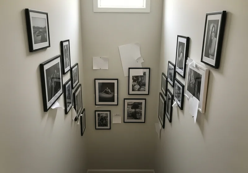 Chaotyczna galeria zdjęć na ścianach klatki schodowej z nierówno rozmieszczonymi ramkami i przyklejonymi kartkami papieru.