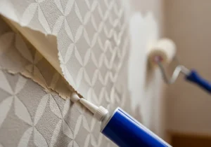 Aplikacja kleju z tubki z precyzyjną końcówką pod krawędź ciężkiej tapety winylowej, w tle wałek do wygładzania powierzchni.