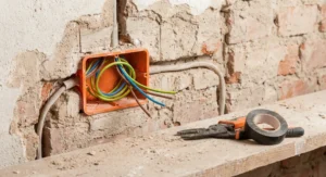 Pomarańczowa puszka elektryczna osadzona w surowej, ceglanej ścianie z wyprowadzonymi, kolorowymi przewodami.