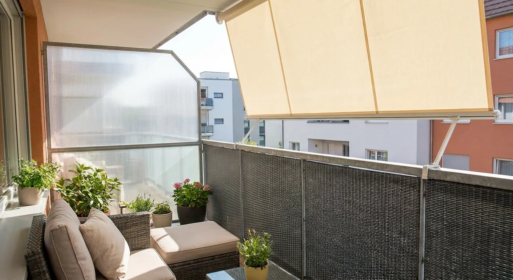 Balkon z osłoną z poliwęglanu, matą technorattanową na barierce i beżową markizą przeciwsłoneczną.