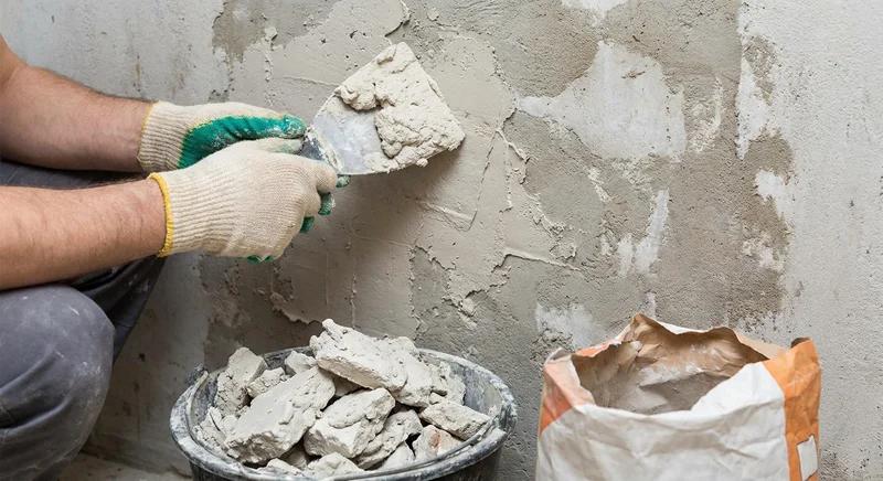Pracownik nakłada gładź na ścianę szpachlą obok wiadra z zaschniętymi bryłami gipsu i otwartego worka.